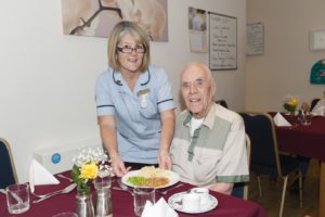 Carer giving plate of food to elderly resident at table